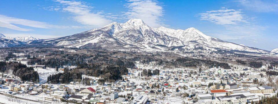 drone view of Myoko Kogen with houses and Mt Myoko covered in snow with a blue sky on the backgound.